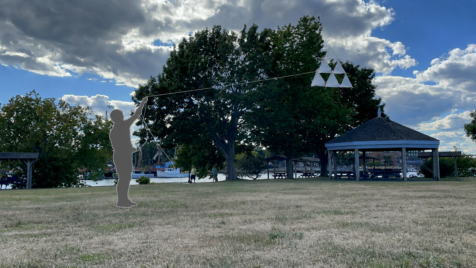 A gray silhouette flies a small white tetrahedral kite in front of a large tree beneath dramatic clouds